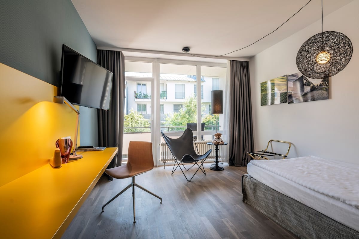 A modern hotel room with a bed, a desk with a chair, a TV mounted on the wall, and a balcony with a view of buildings and greenery.