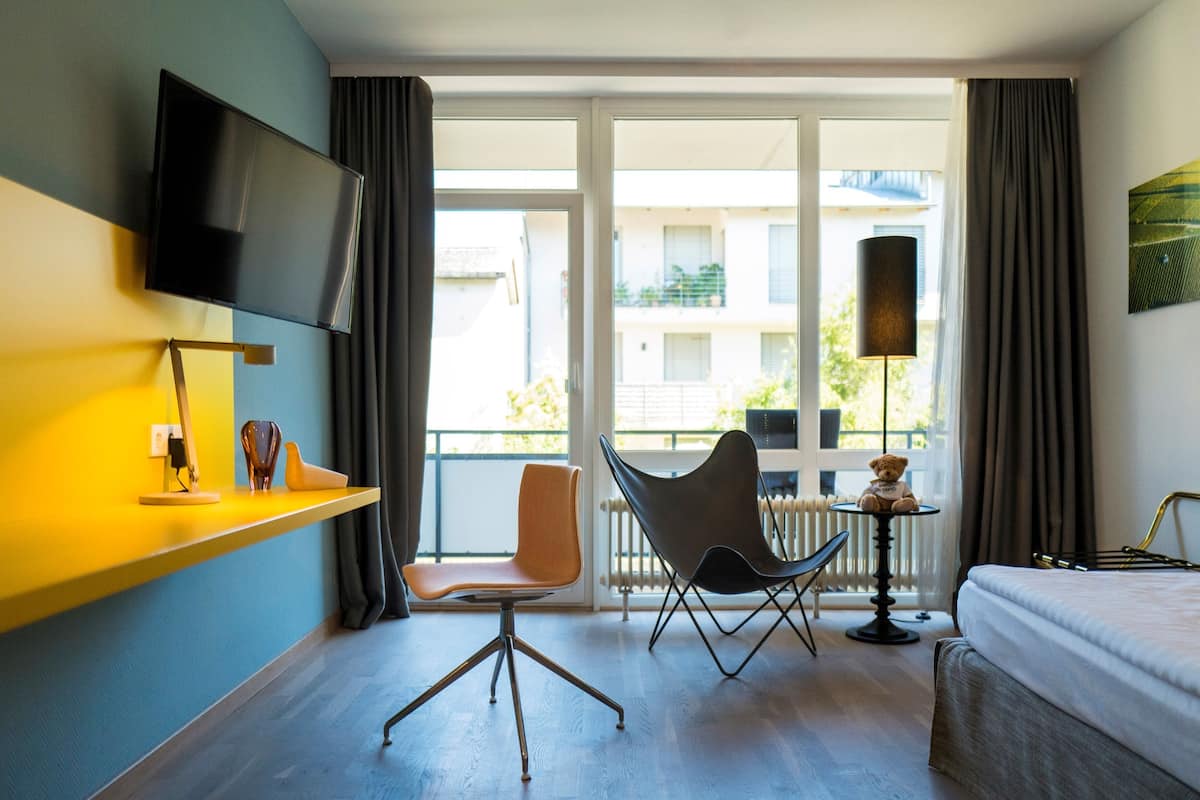 A hotel room with a bed, a desk with a chair, a TV mounted on the wall, and a view of the outside balcony.