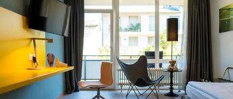 Habitación de hotel con cama, escritorio con silla, televisor montado en la pared y vistas al balcón exterior.