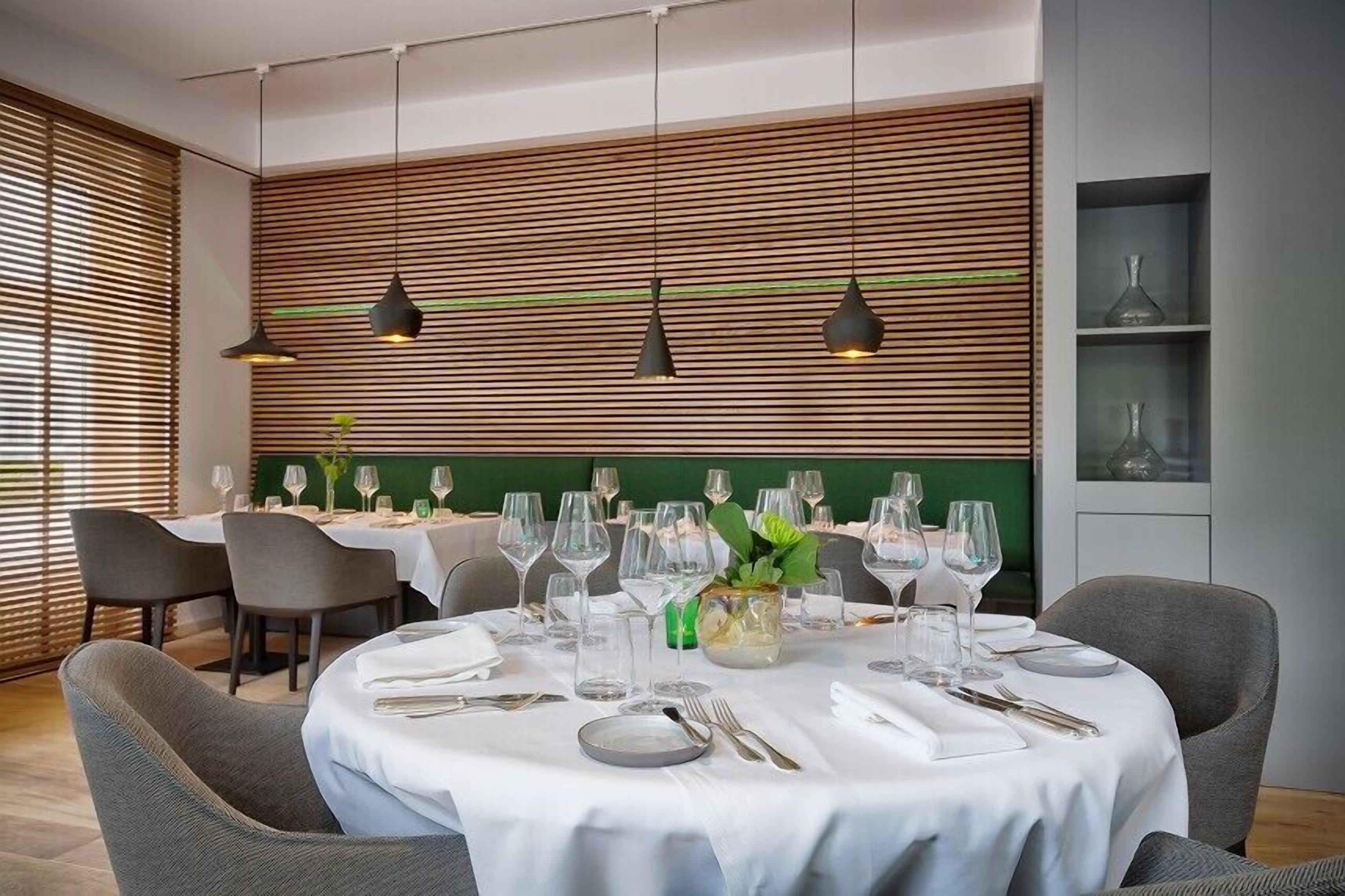 Un comedor con mesas redondas dispuestas para una comida, con una pared verde con listones de madera horizontales e iluminación colgante.
