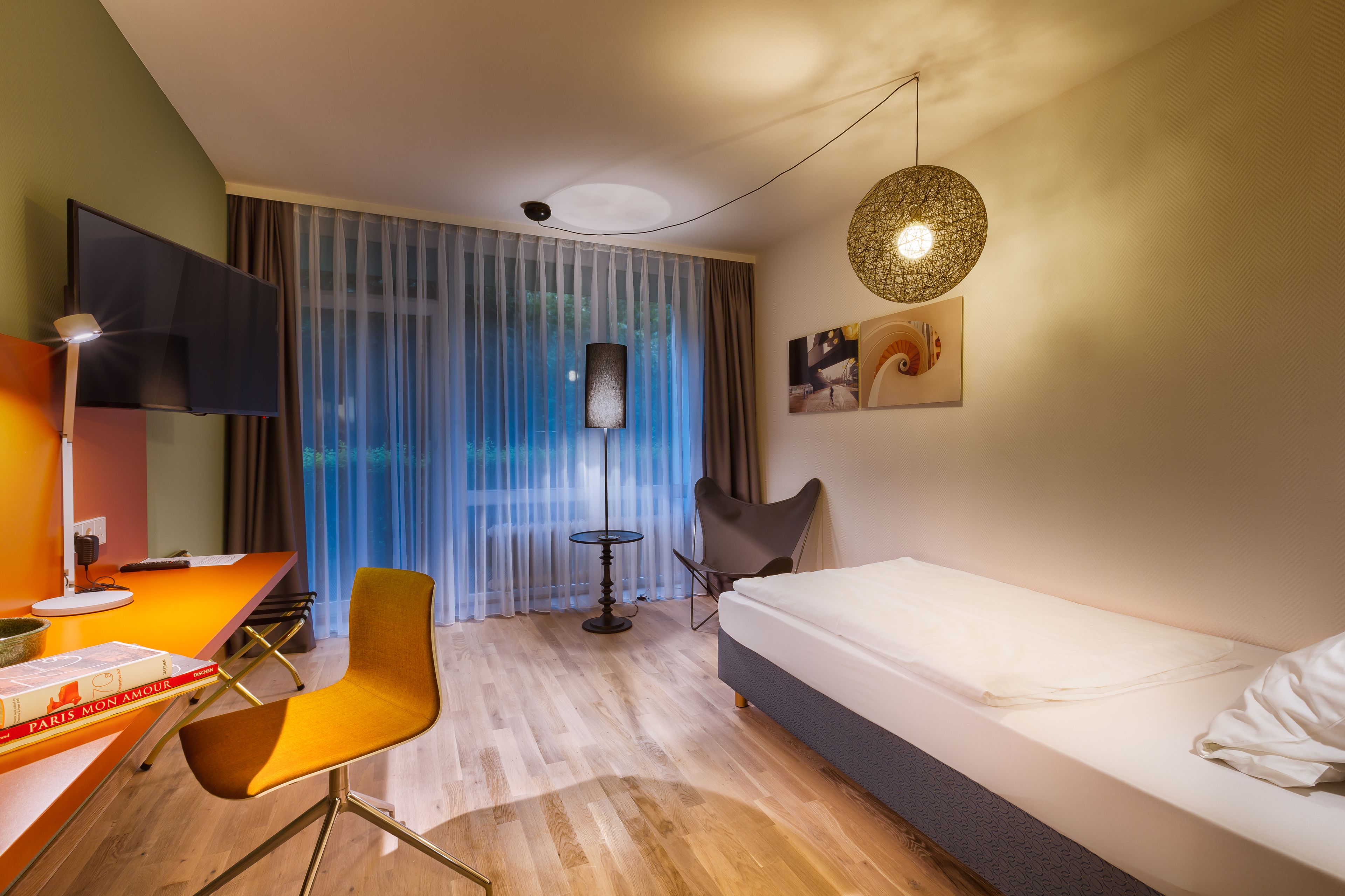Ein modernes Hotelzimmer mit einem Bett, einem Schreibtisch mit Stuhl, einem Fernseher, einer Lampe und Blick ins Freie durch transparente Vorhänge.