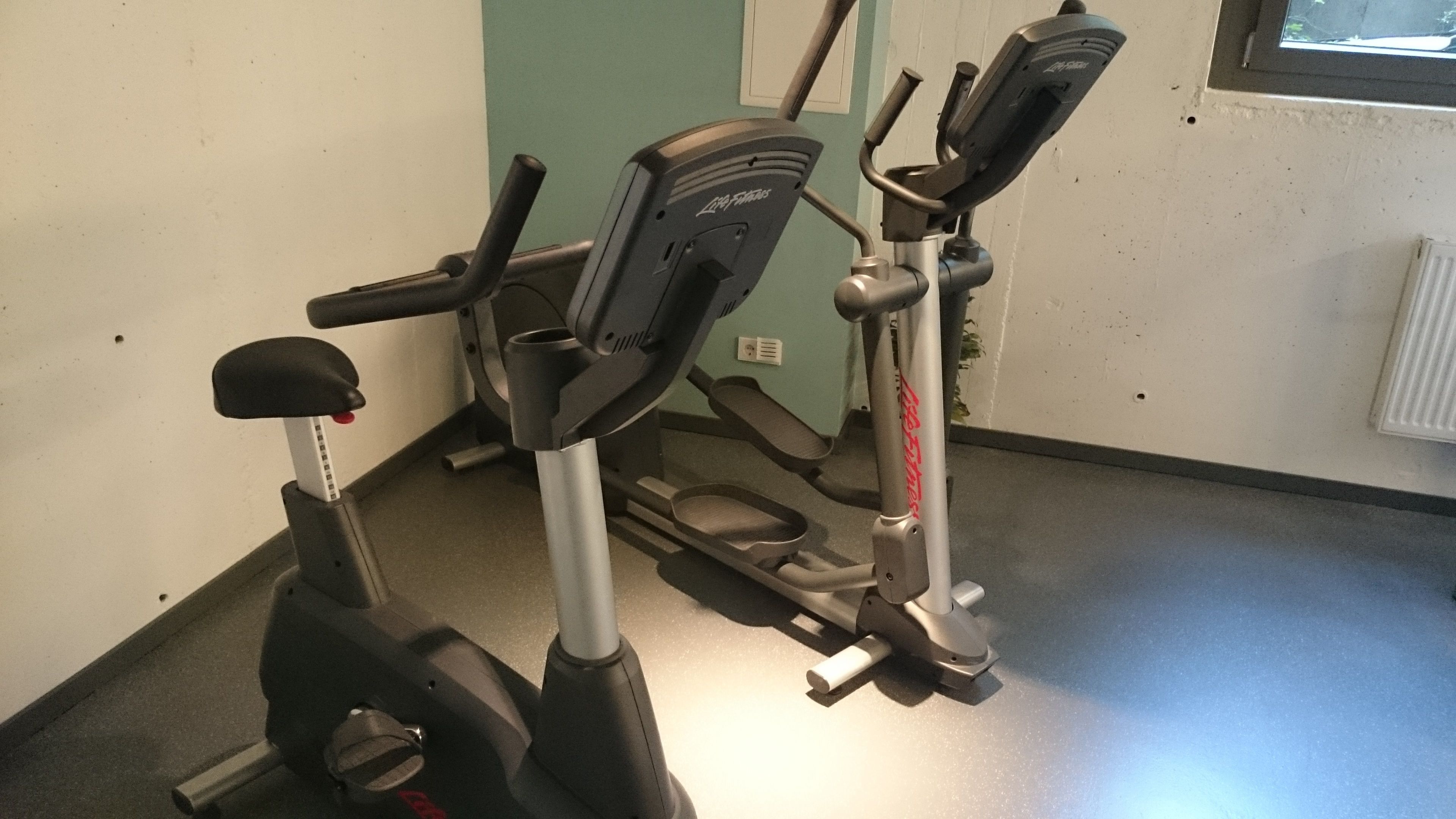 A stationary bike and a treadmill in a room with a plain wall and a window.