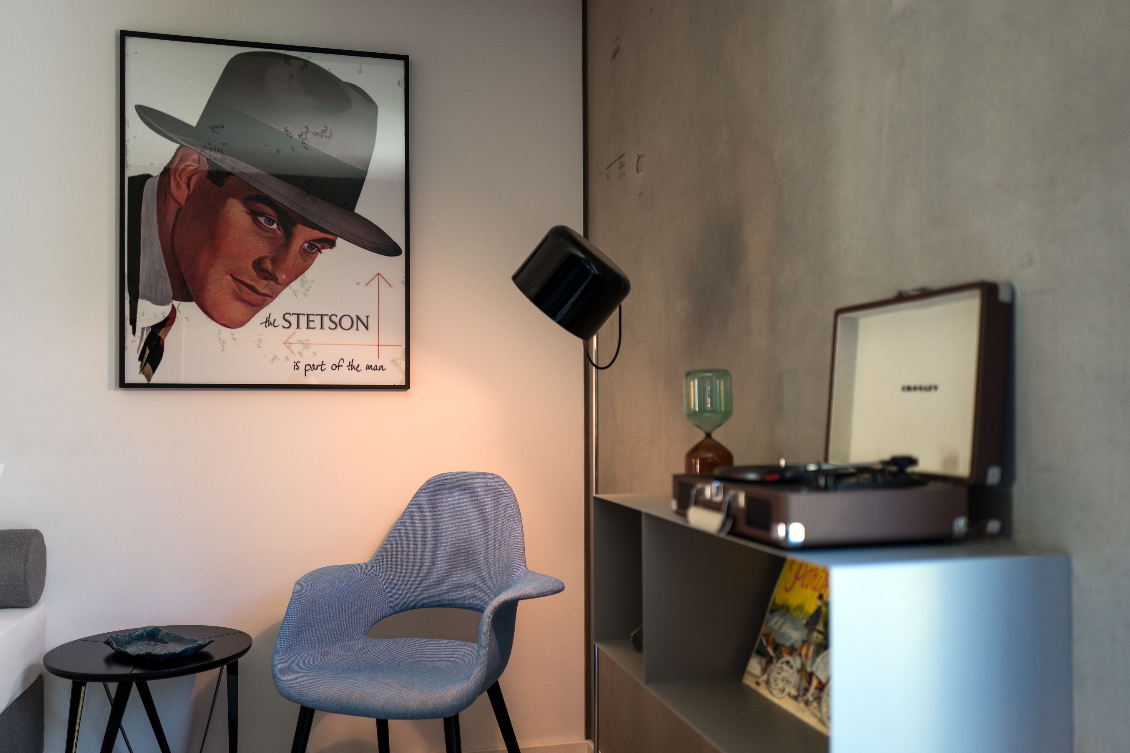 A room with a framed poster, a blue chair, a small table, a turntable, and a lamp.