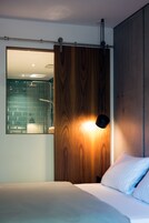 Un dormitorio moderno con un armario de madera, una cama con ropa de cama blanca y un baño visible a través de una puerta de cristal.