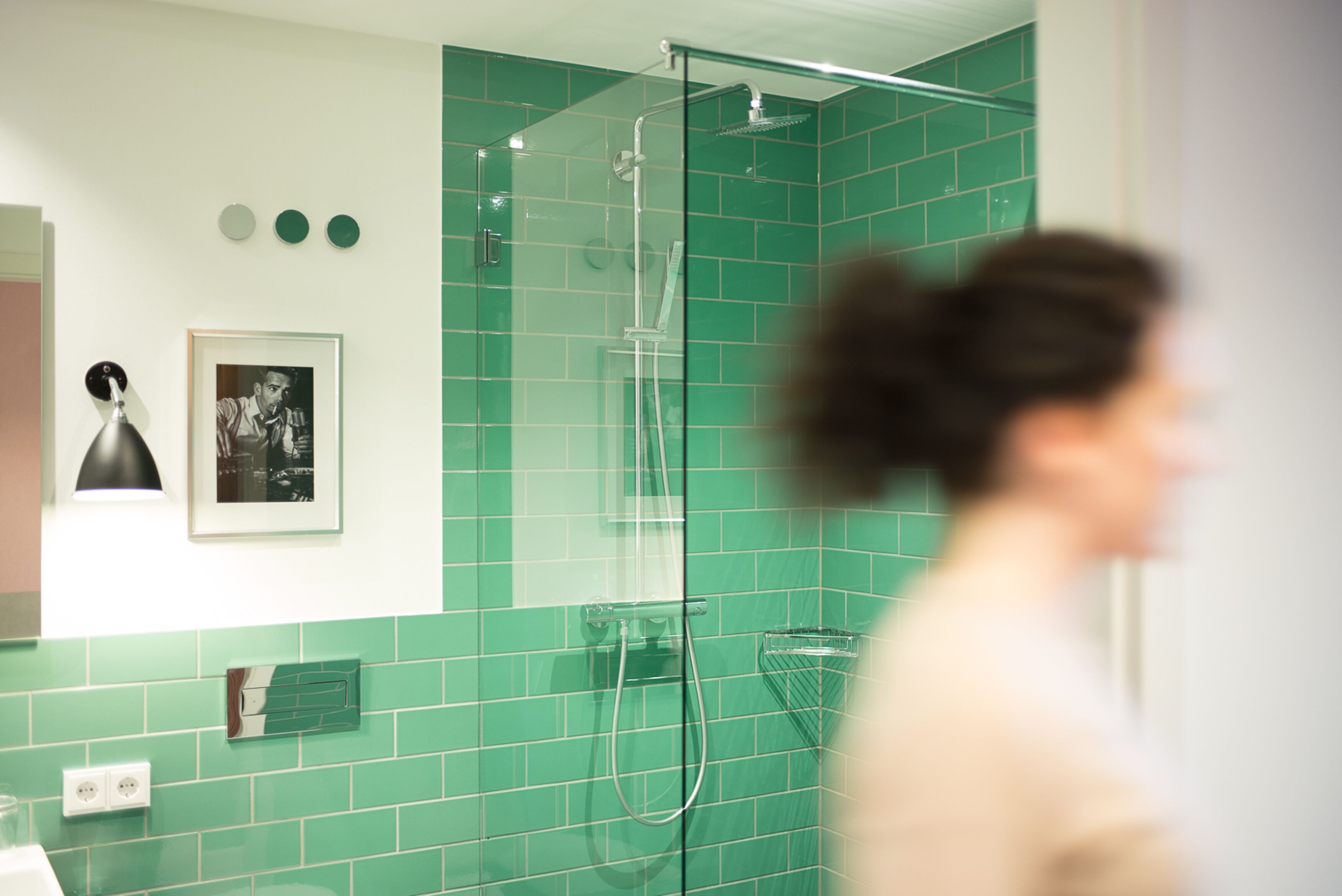 A bathroom with green tiles, a glass shower enclosure, a framed picture on the wall, and a towel rack.