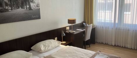 Een hotelkamer met twee bedden, een bureau, een stoel, een lamp en een groot raam met gordijnen.
