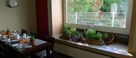 A dining area with a table set for four, a window with a view of greenery, and a shelf with potted plants.