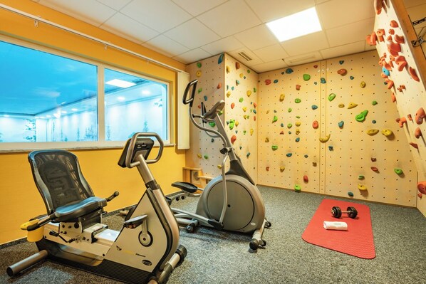 A gym with exercise equipment, a climbing wall, and a window with a view. - Berghotel Jaegerhof (Isny im Allgaeu)