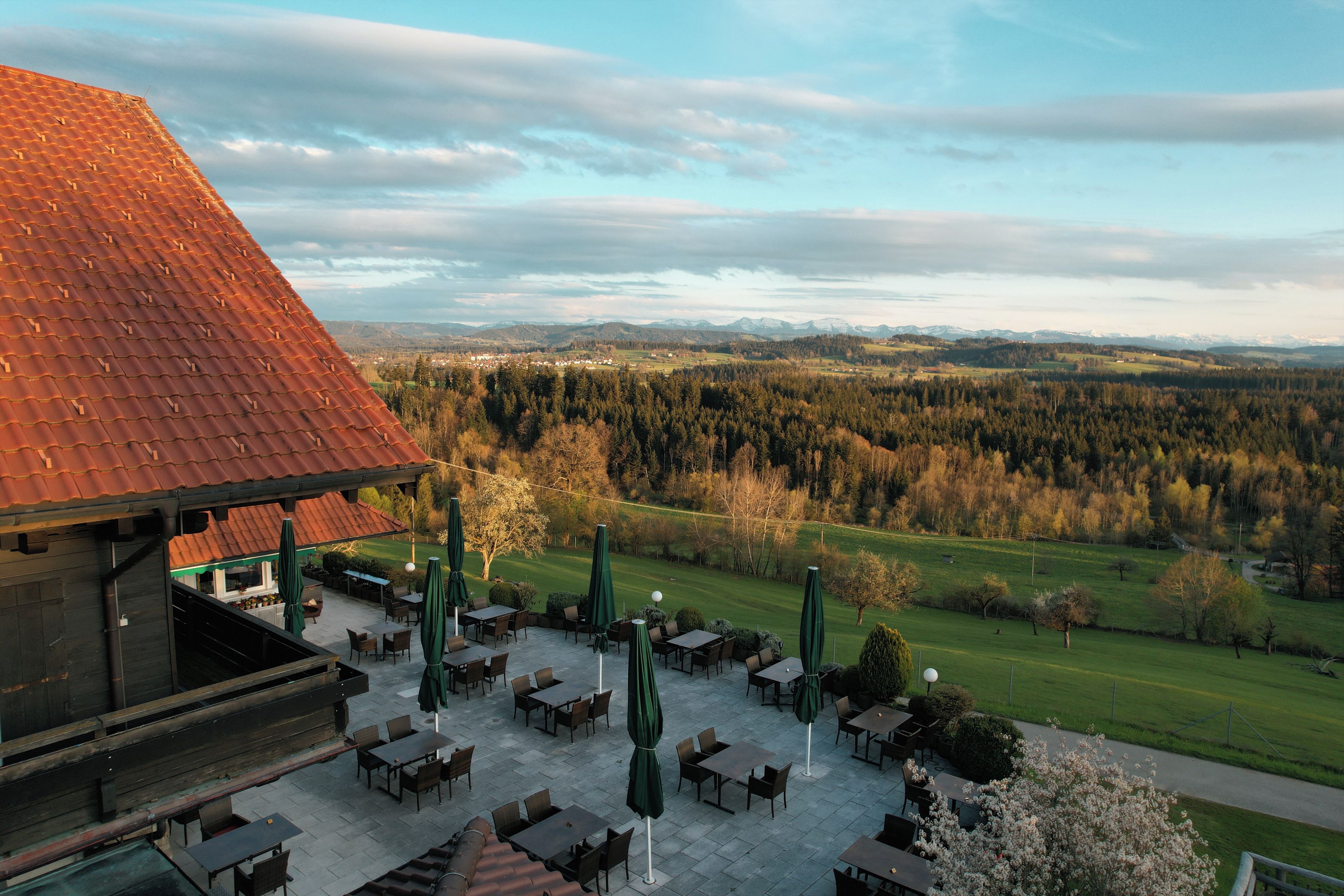 A terrace with outdoor seating, a view of a mountainous landscape, and a building with a red roof.