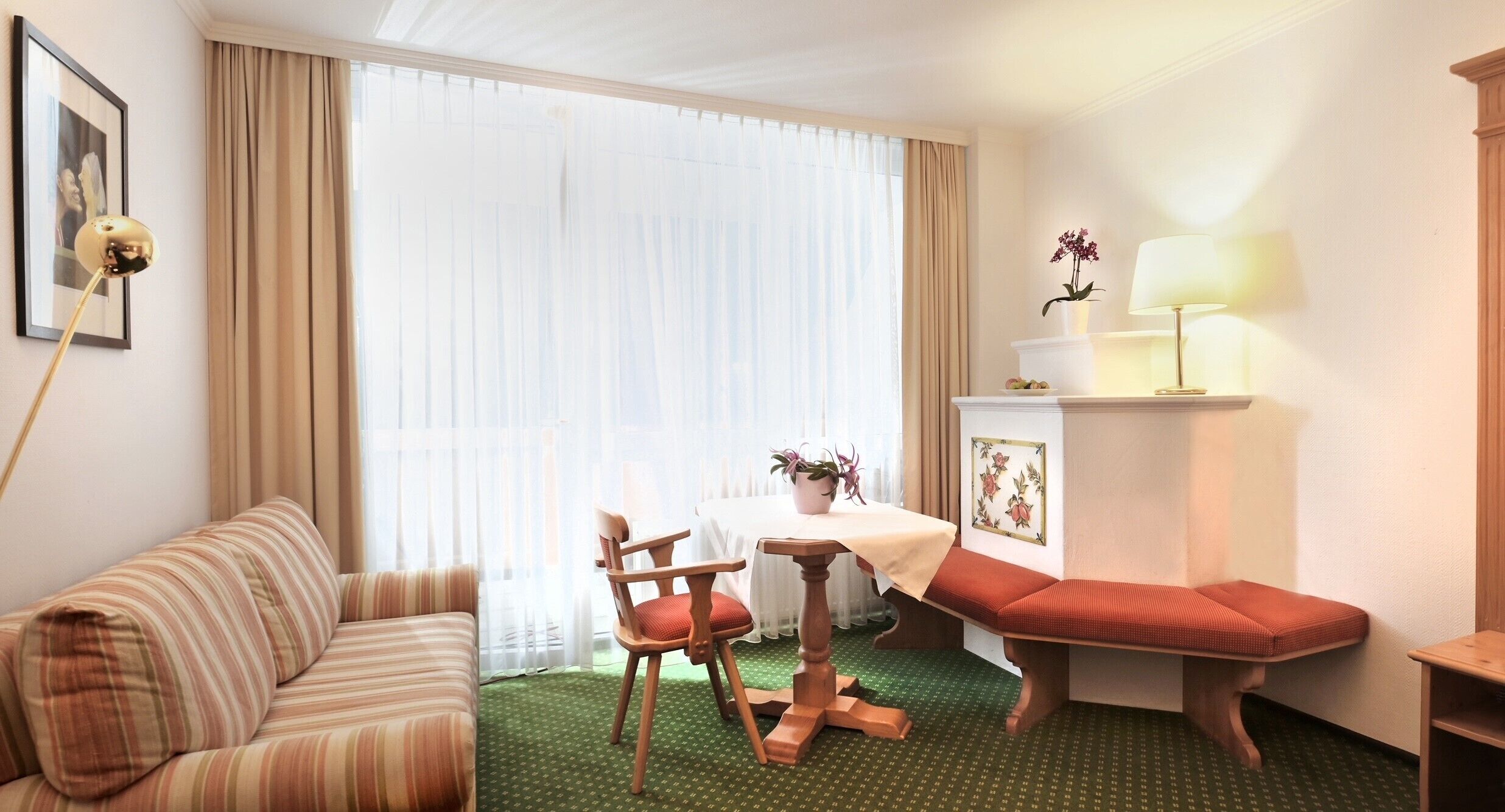 A hotel room with a striped sofa, a wooden chair, a small table with a white tablecloth, a red bench, a lamp, and a potted plant.