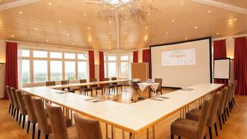Un salón de conferencias con una larga mesa, sillas, una pantalla de proyector y vistas al exterior a través de amplios ventanales.