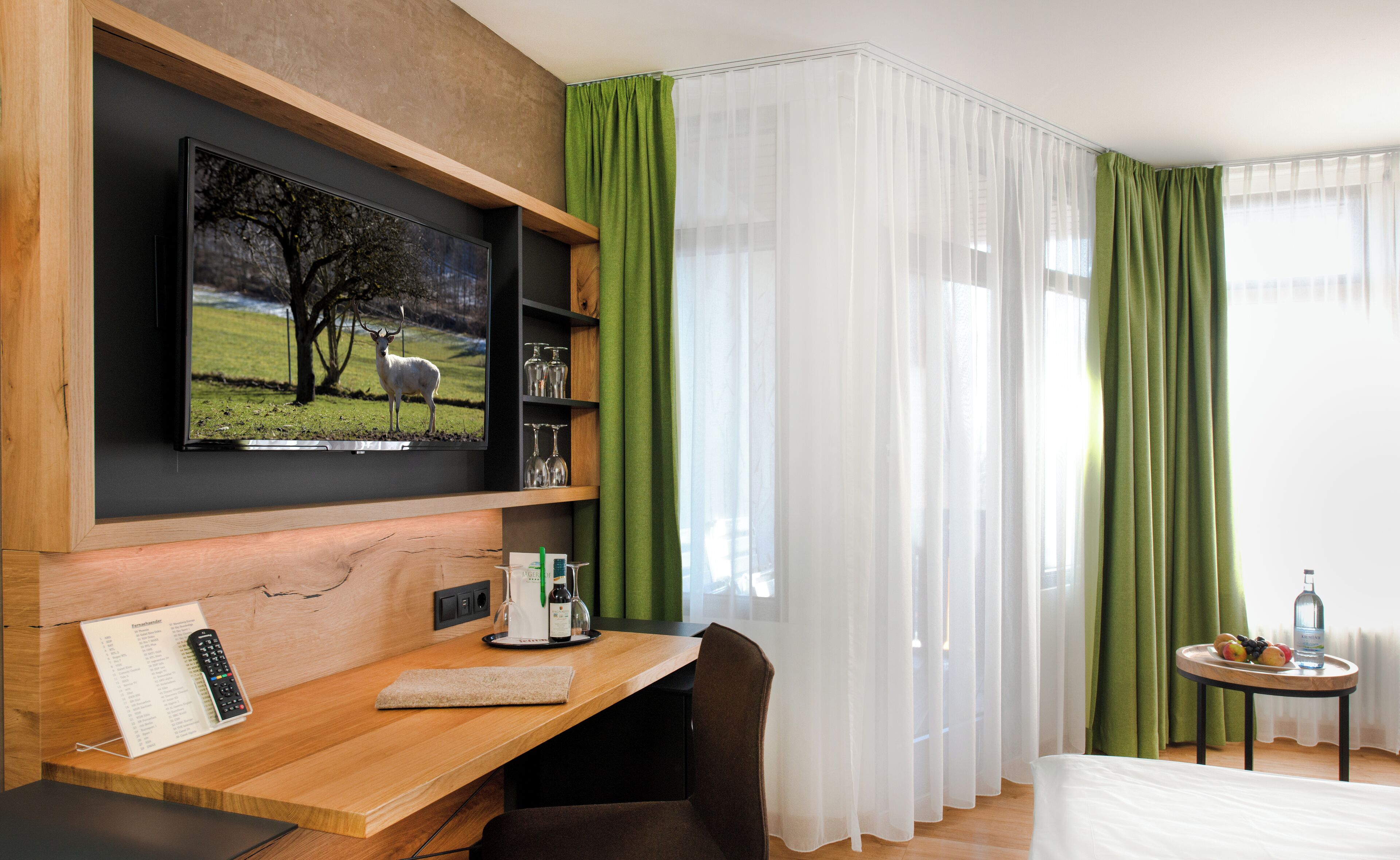 A hotel room with a wooden desk, a flat-screen TV mounted on the wall, a bed, a small table with a bottle and fruits, and green curtains.