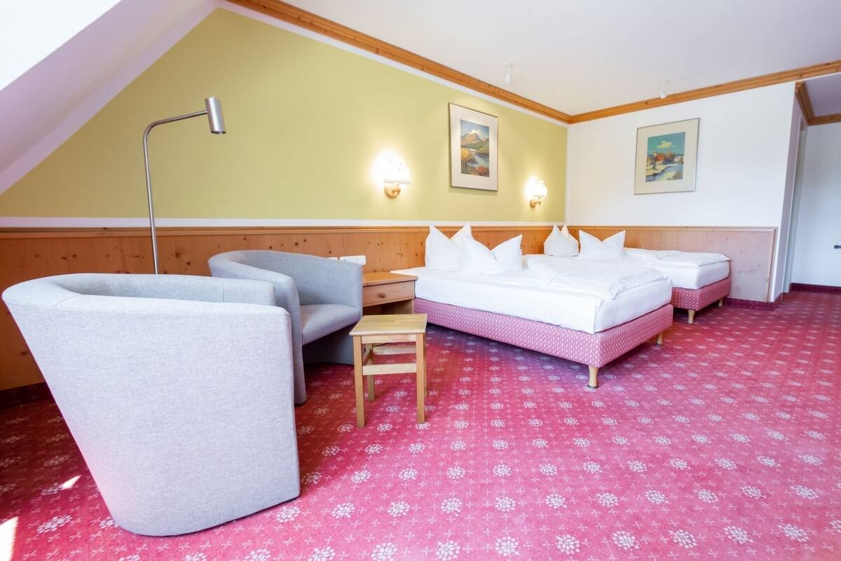 A room with a sloped ceiling, two beds with white bedding, a grey armchair, a small wooden table, and a red patterned carpet.