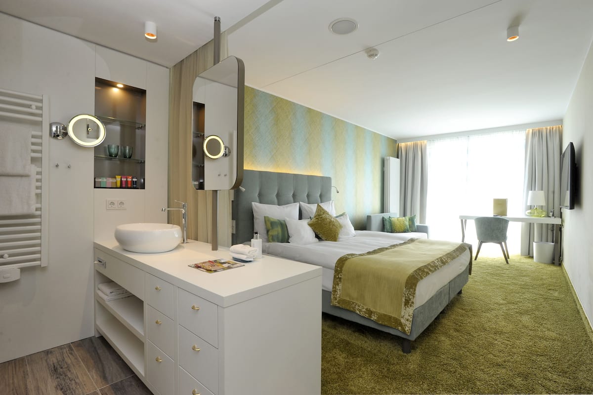 A modern hotel room with a large bed, a bedside table, a built-in vanity with a mirror, and a desk with a chair.
