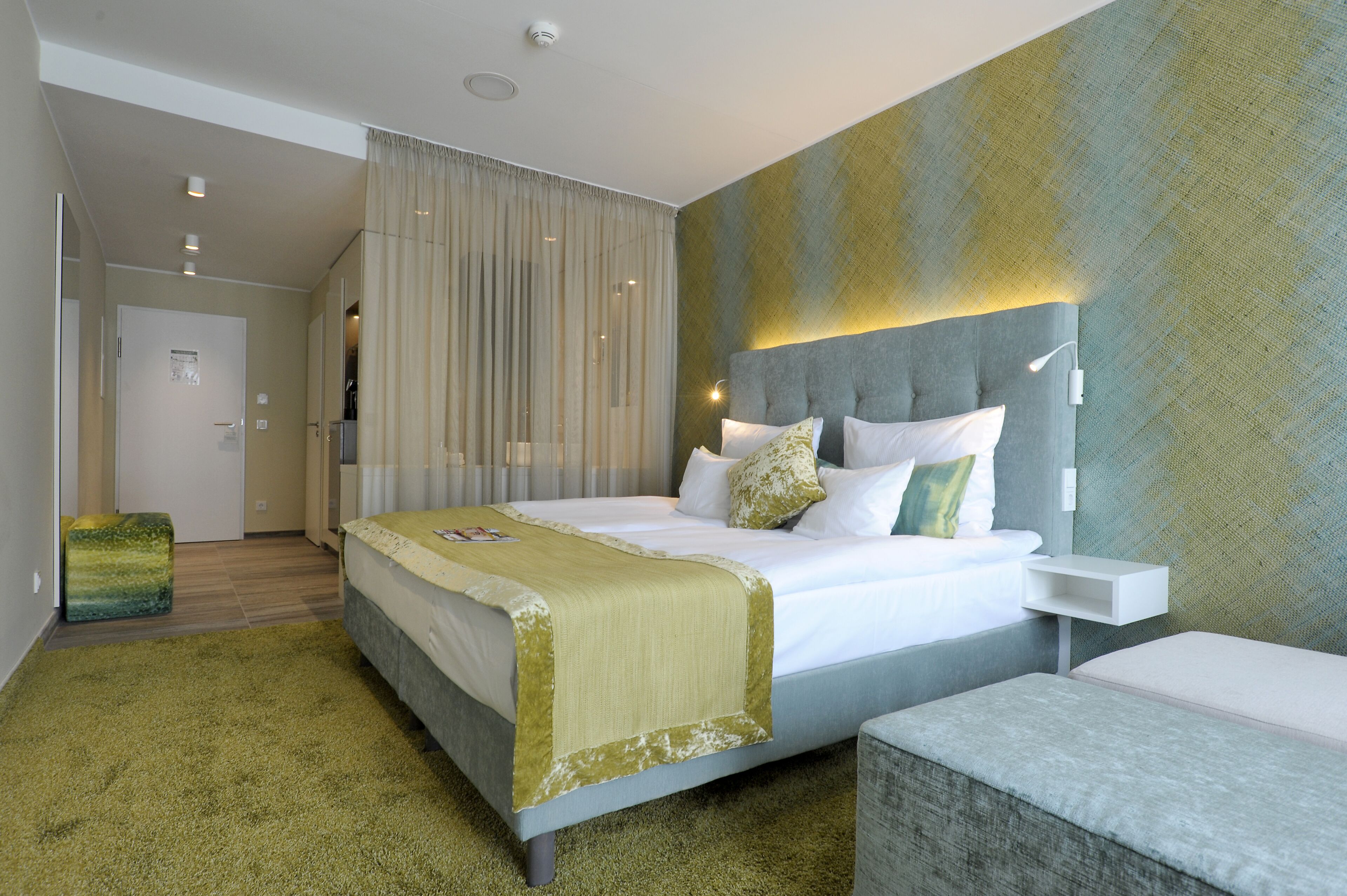 A modern hotel room with a large bed, a bedside table, a green ottoman, and a view of the hallway.