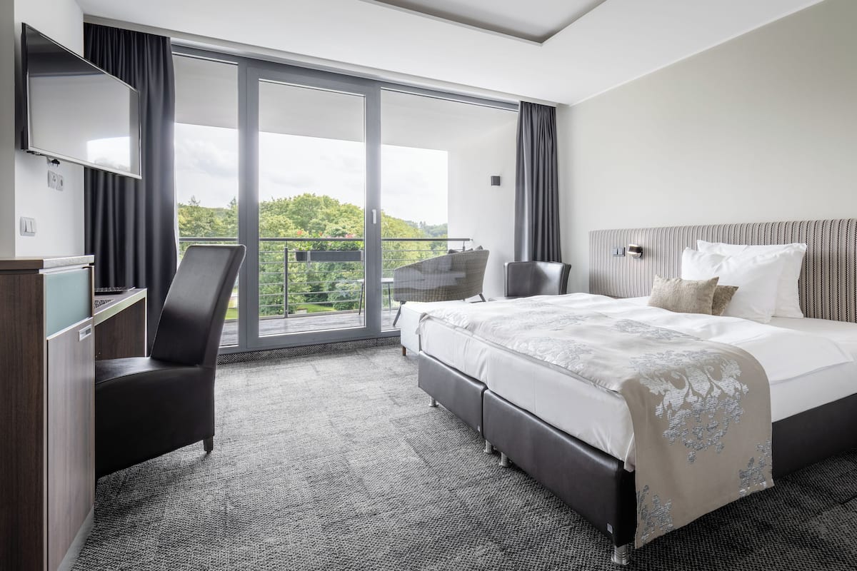 A modern hotel room with a large bed, a desk, and a balcony with a view of greenery.