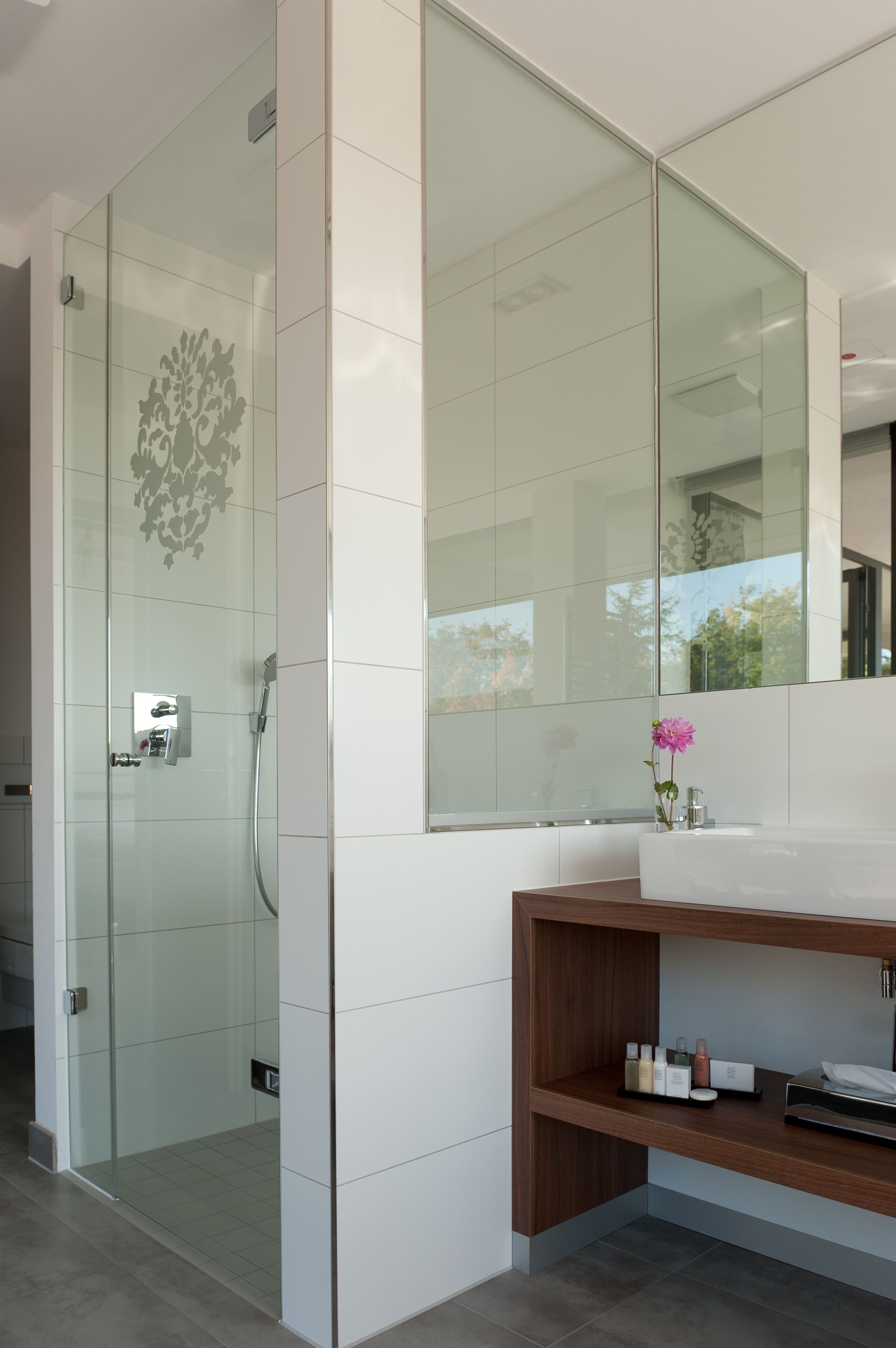 Un baño moderno con cabina de ducha de vidrio, un espejo grande y un tocador de madera con artículos de aseo.