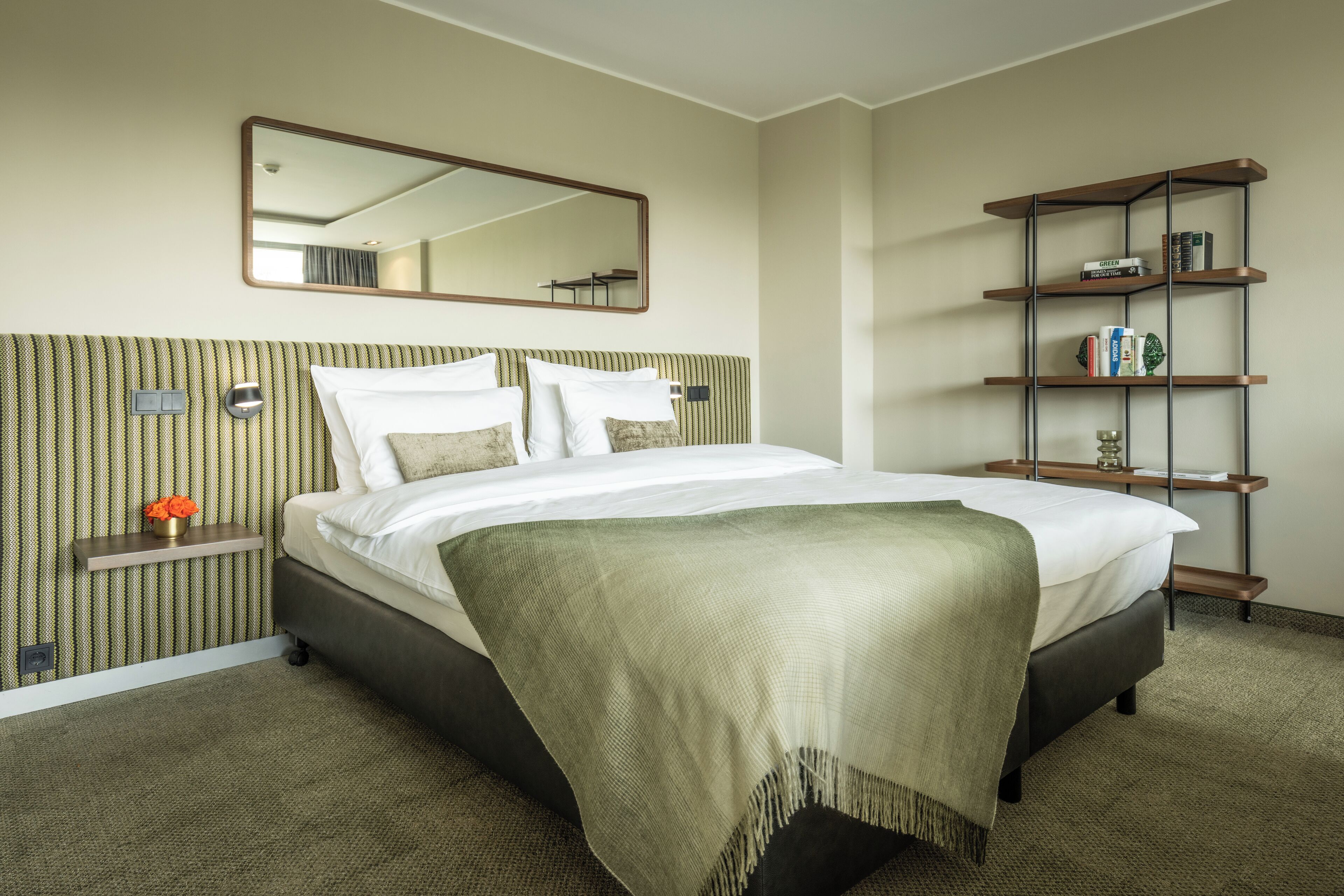 A hotel room with a bed, a mirror, a shelf with books, and a carpeted floor.