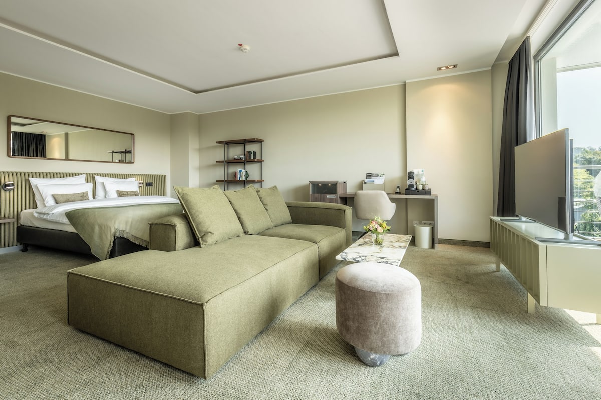 A modern hotel room with a large bed, a sofa, a small ottoman, a desk, and a television.