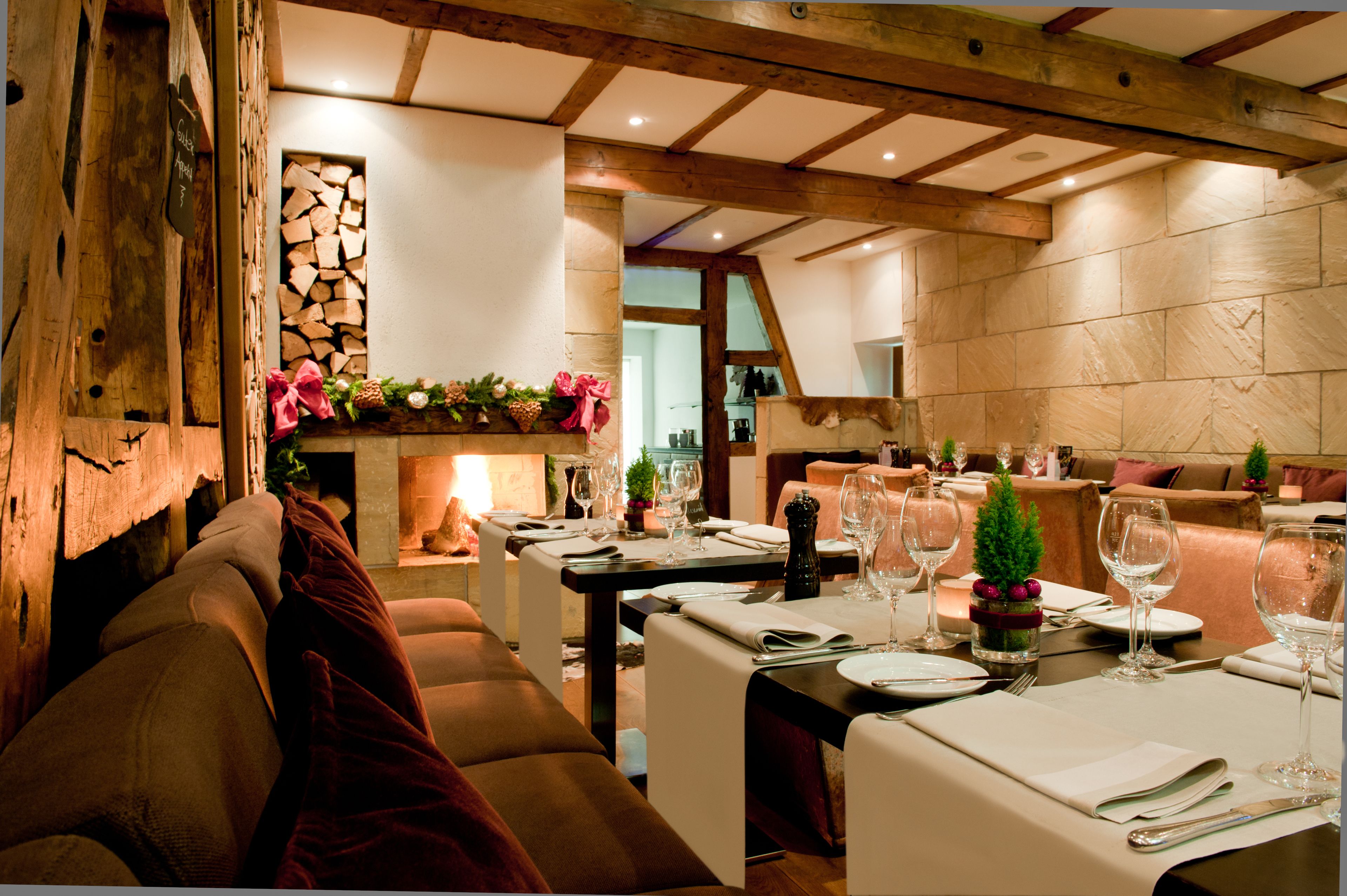 Un restaurante con vigas de madera, paredes de piedra y una zona de estar acogedora. Las mesas están dispuestas con manteles blancos, platos y copas de vino.