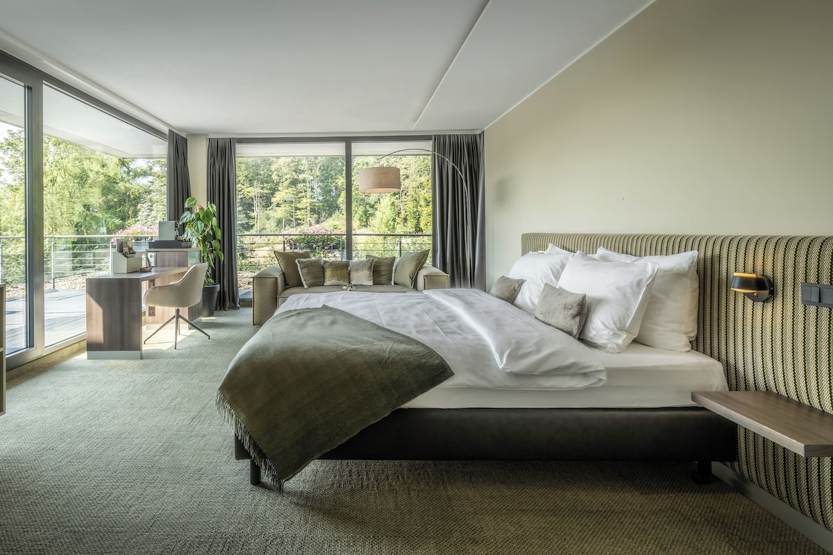 A modern bedroom with a large bed, a desk, and a balcony with a view of trees.
