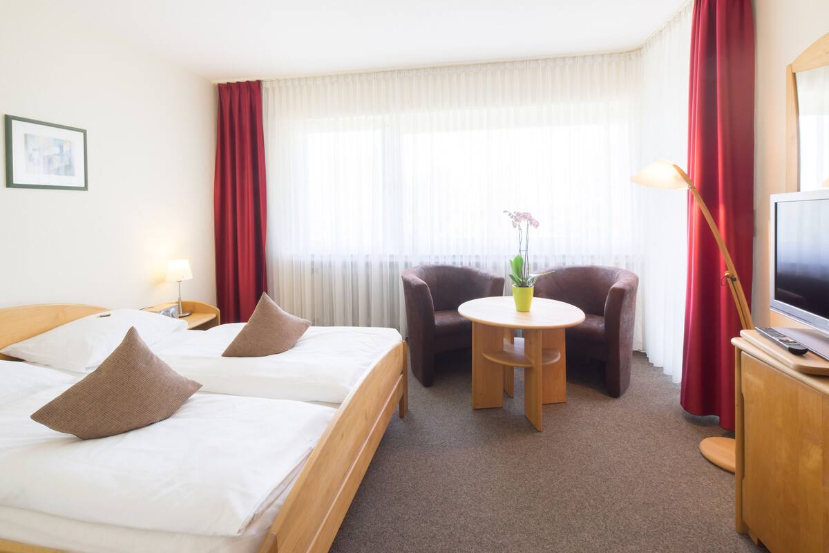 A hotel room with two beds, a small round table with a potted plant, a chair, a desk with a TV, and a window with curtains.