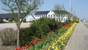 A building with a white facade and dark roof, surrounded by a garden with red and yellow flowers, and a clear sky. - Den Gamle Grænsekro (Christiansfeld)