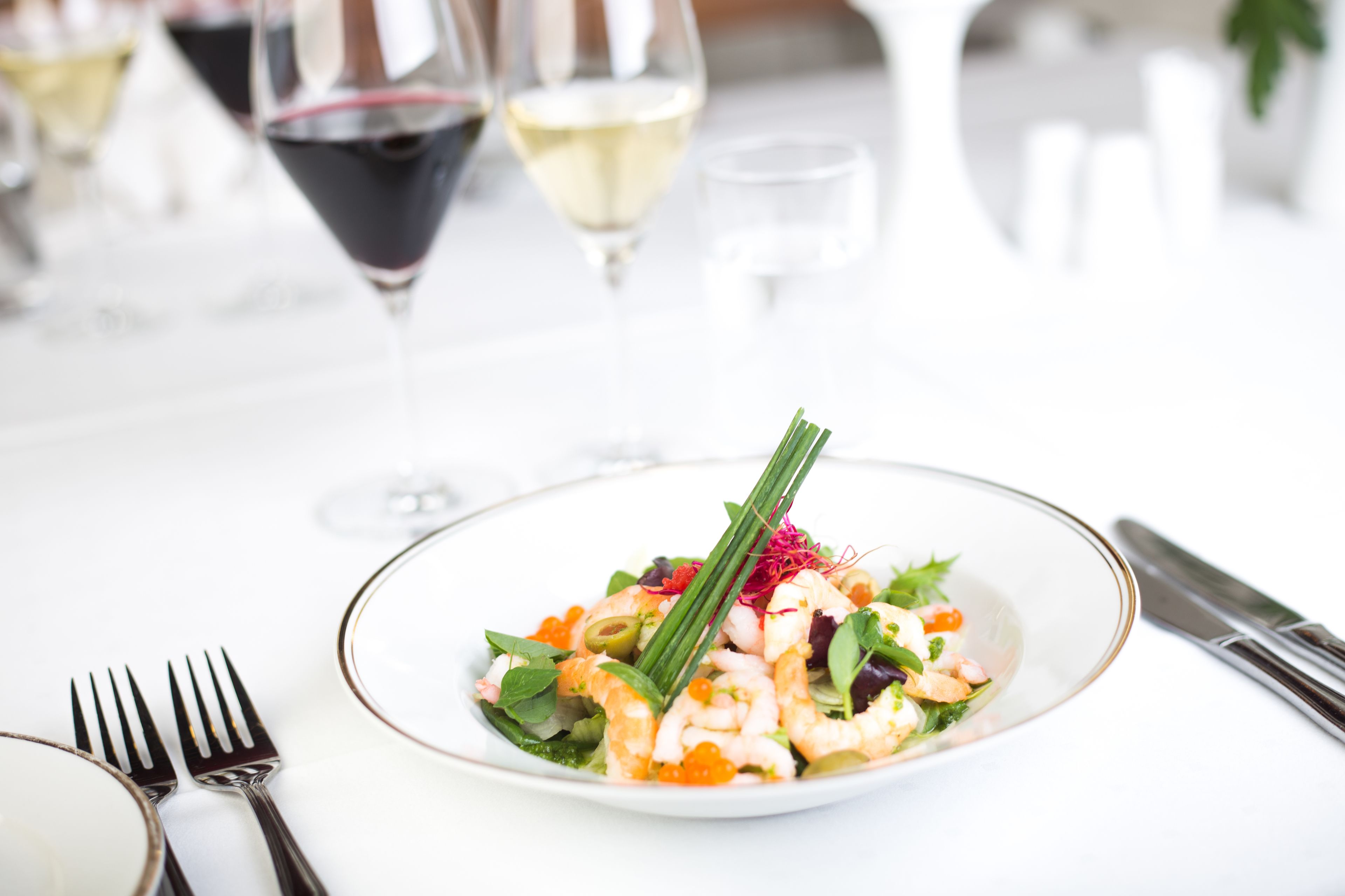 A plate of shrimp salad with a garnish, accompanied by wine glasses and cutlery on a white table.