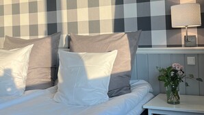 A bedroom with a checkered wall, a bed with white and grey bedding, a bedside table with a lamp, and a vase with flowers. - Den Gamle Grænsekro (Christiansfeld)