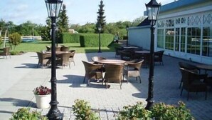 An outdoor patio area with wicker furniture, a white building with patterned windows, and a hedge in the background. - Den Gamle Grænsekro (Christiansfeld)