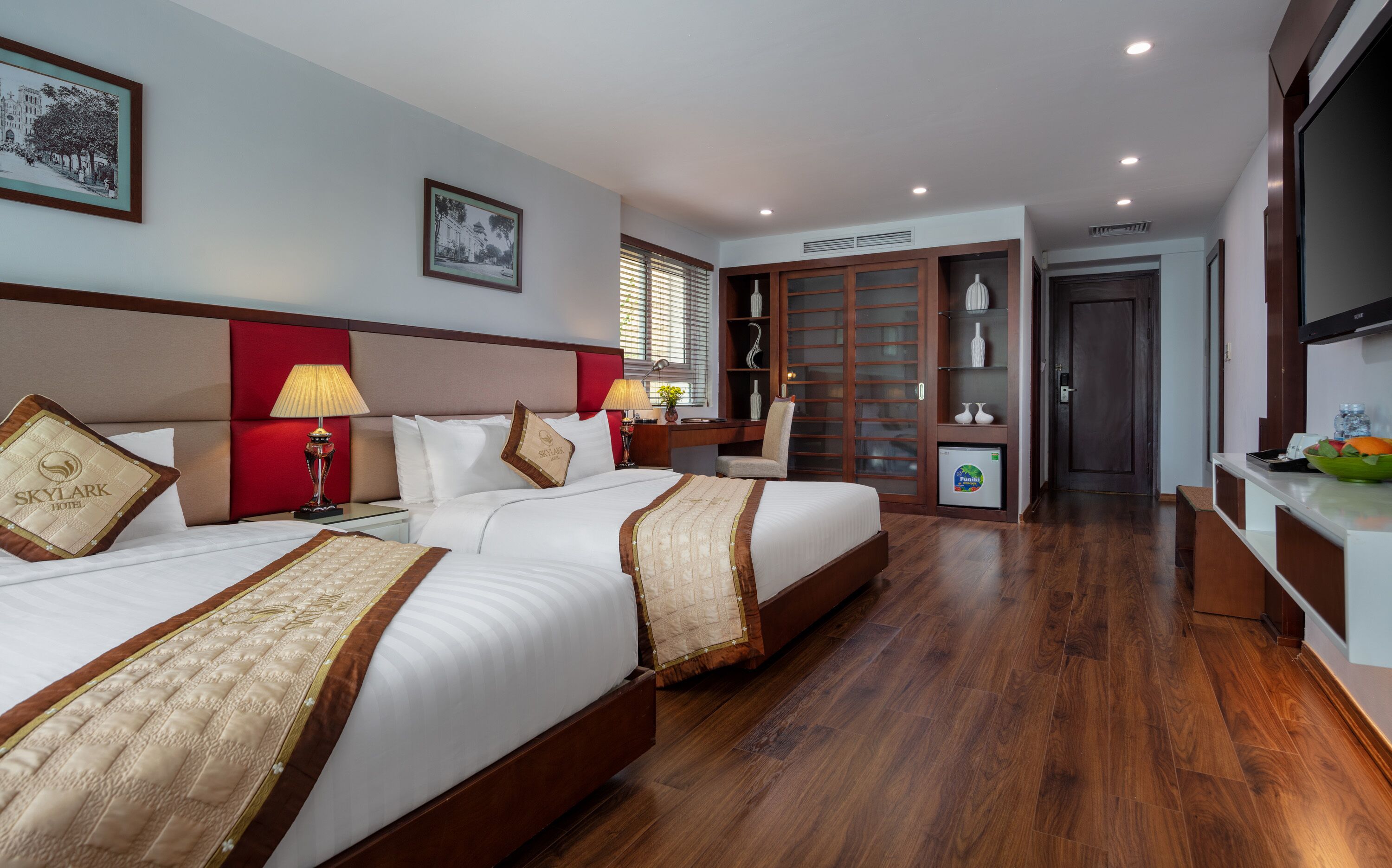 A hotel room with two beds, a desk, a chair, a wardrobe, a TV, and a mini-fridge.