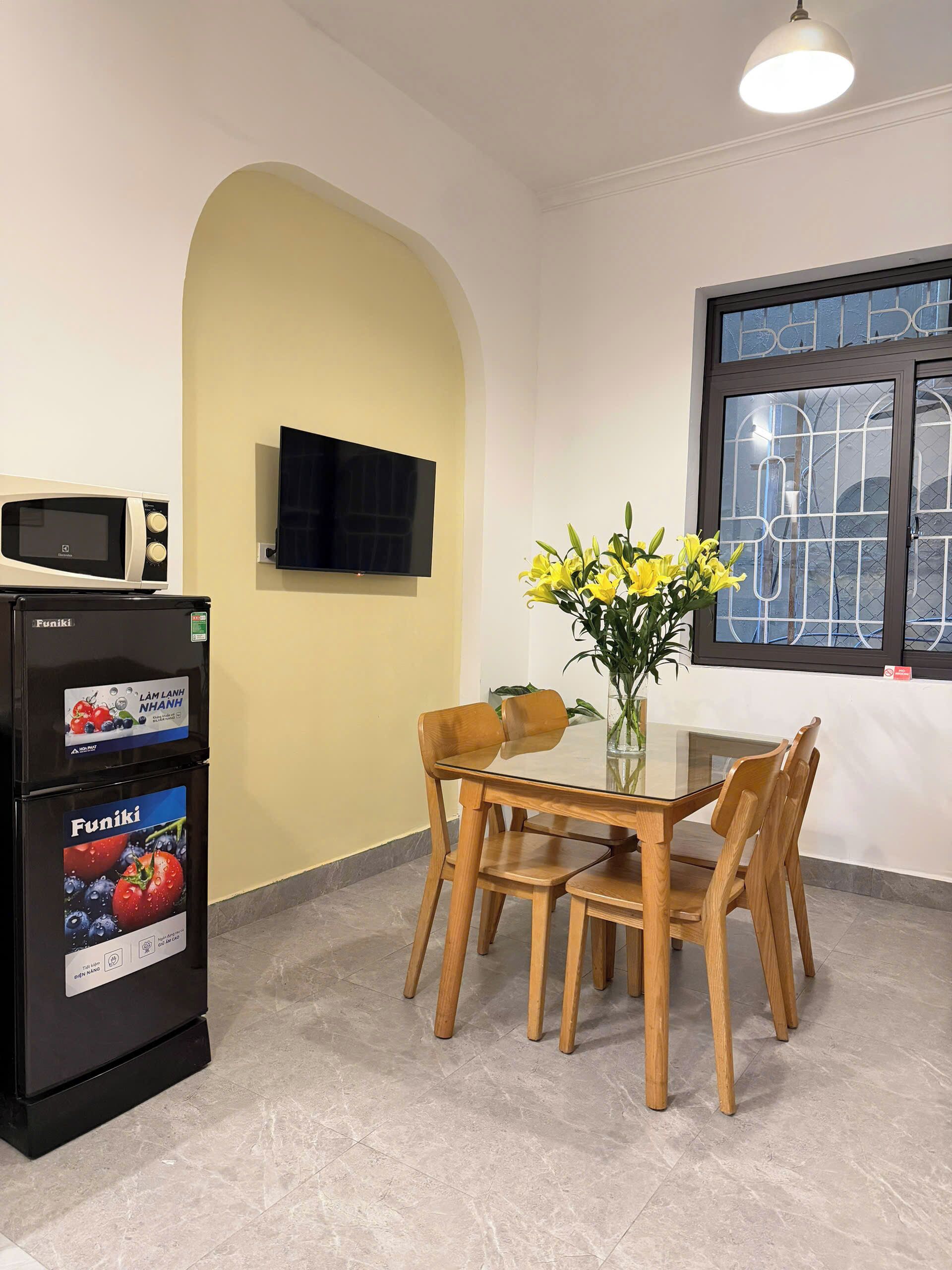 A dining area with a table and chairs, a wall-mounted TV, a refrigerator with product labels, and a window with a metal grille.