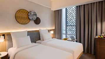 A hotel room with two beds, a small table with a laptop, a wicker side table, and a window with curtains.