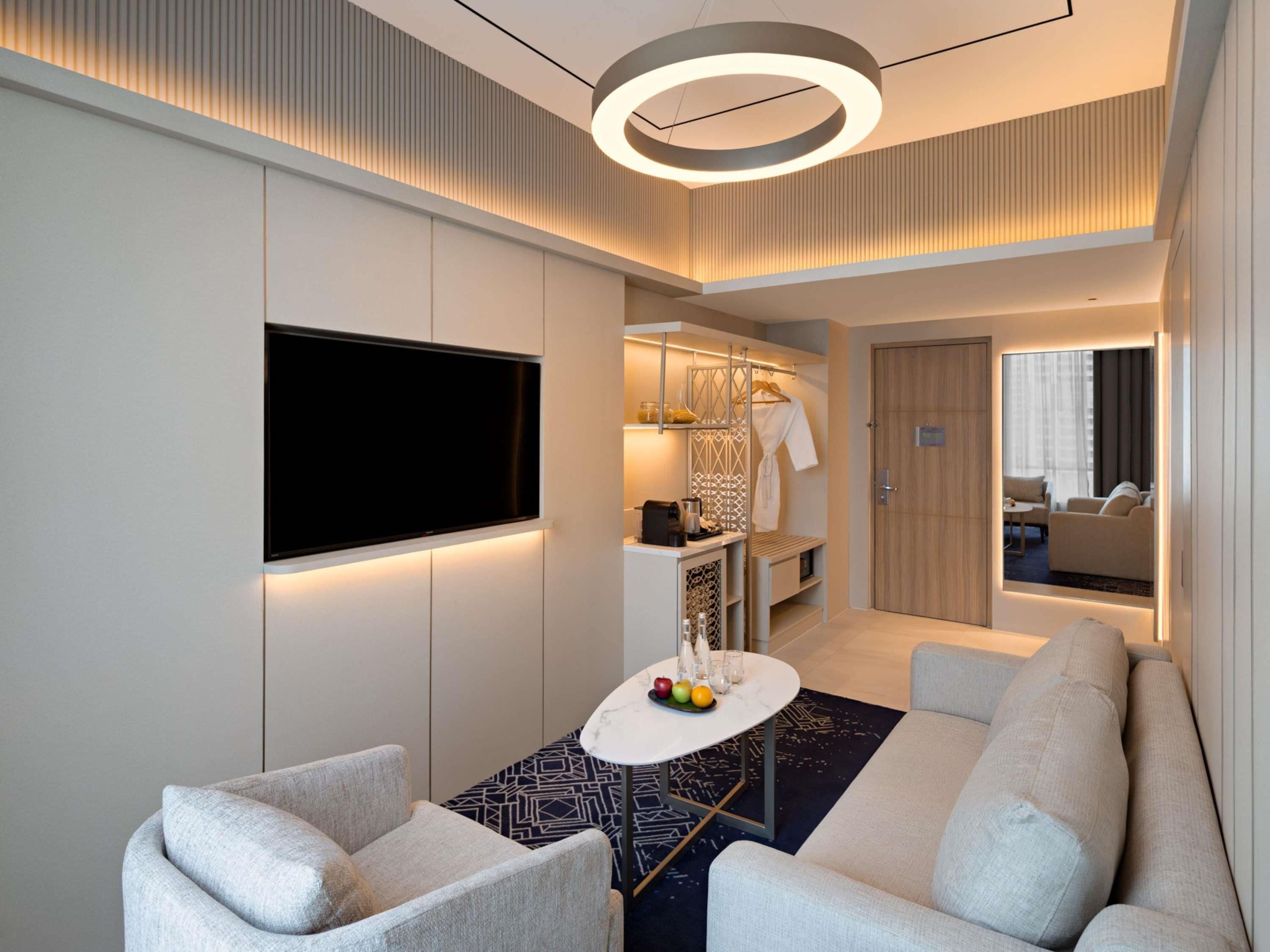 A modern hotel room with a flat-screen TV, a round ceiling light, a seating area with a small table, and a view into the living area.