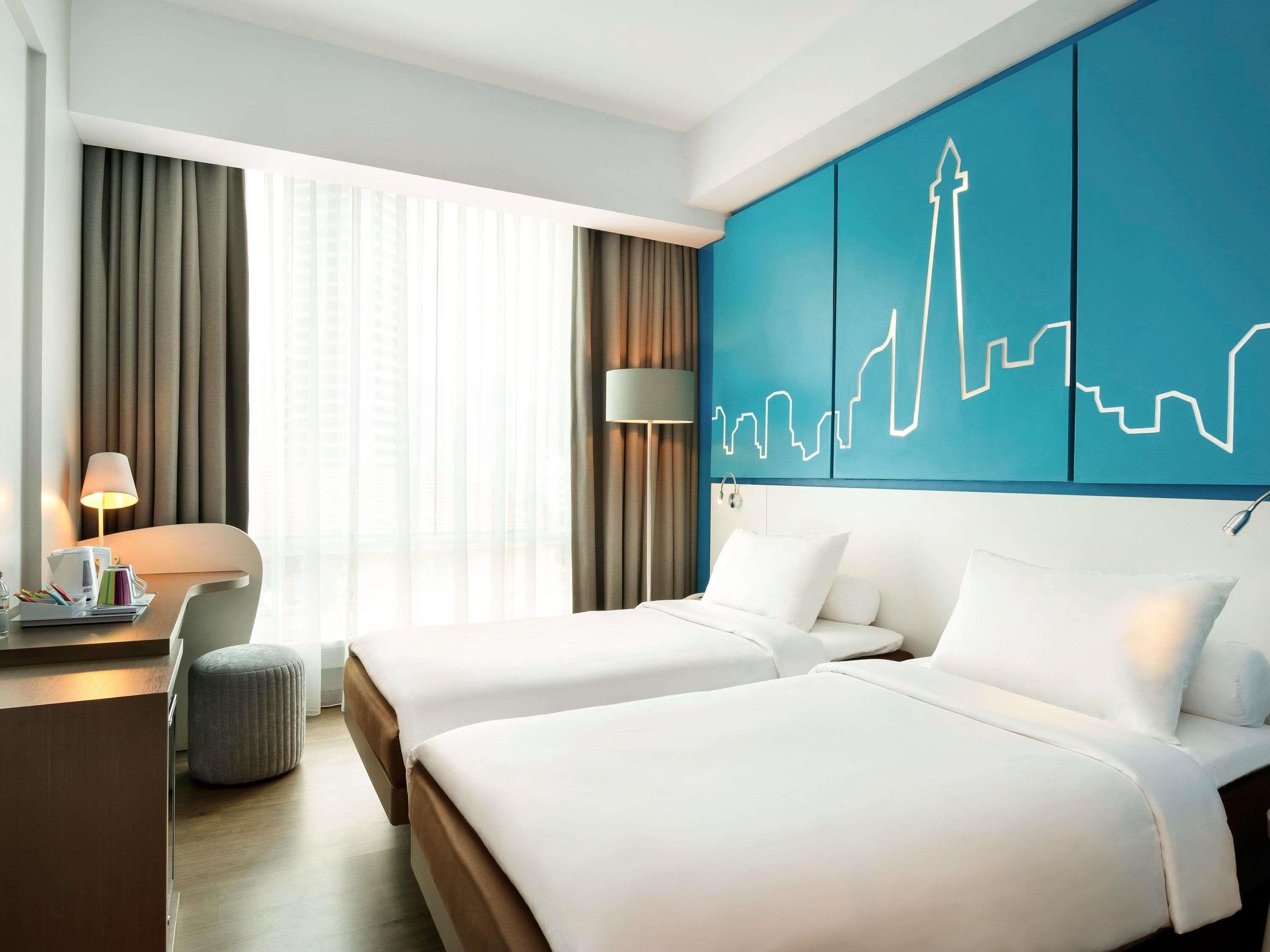 Une chambre d’hôtel avec deux lits, un bureau, une lampe et un mur décoré d’un motif représentant le panorama urbain.