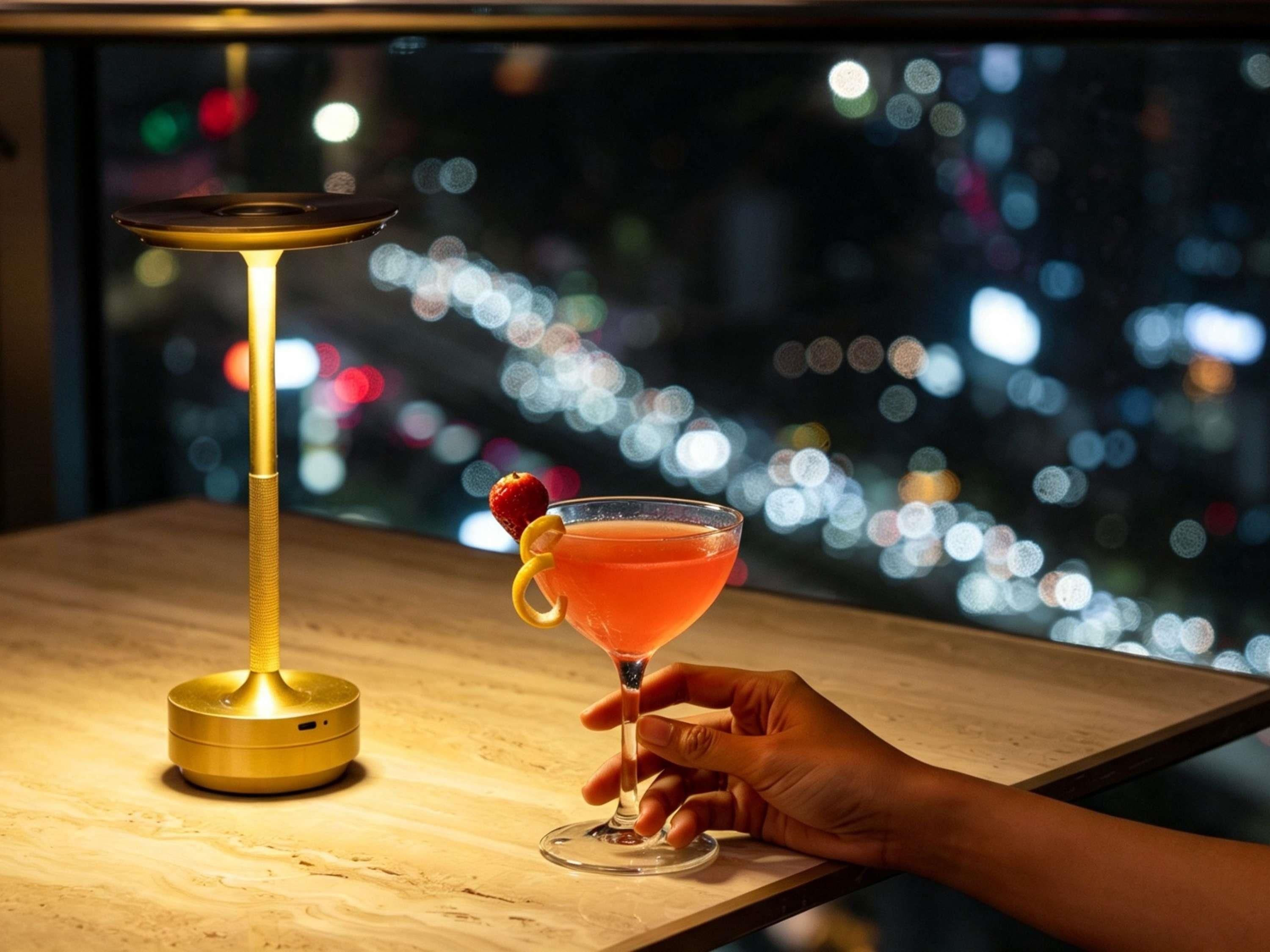 A cocktail in a martini glass with a strawberry garnish, held by a hand, next to a golden cocktail shaker on a wooden surface with a cityscape view in the background.