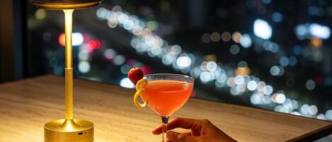 Un cóctel en una copa de martini adornado con una fresa, sostenido por una mano, junto a un coctelero dorado sobre una superficie de madera con vista a un paisaje urbano de fondo.