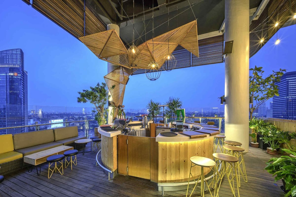 A rooftop bar with a wooden bar counter, seating, and potted plants.