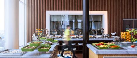 A buffet area with various food stations, a person in a yellow vest, and a large window in the background.