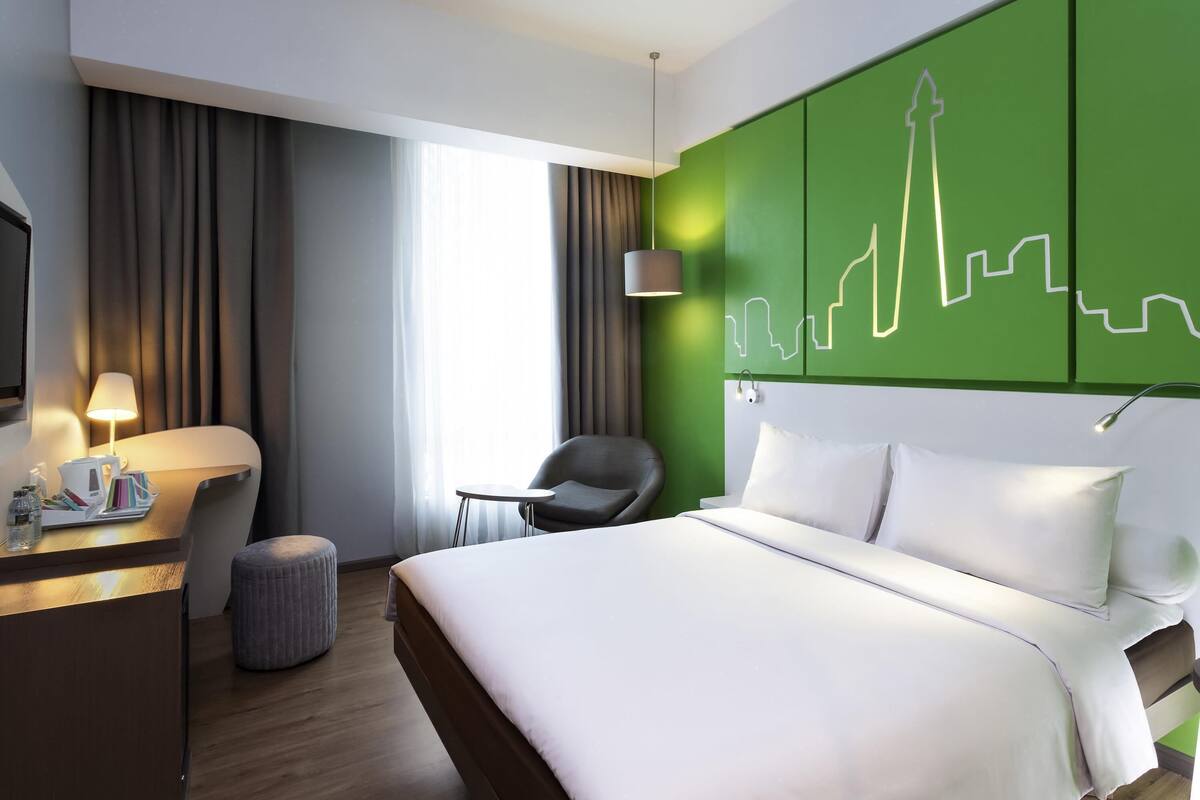 A modern hotel room with a bed, a desk, a chair, and a green accent wall.