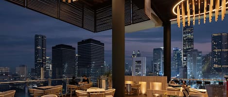 Un área de comedor al aire libre con mesas y sillas, una terraza de madera y vistas a un horizonte urbano.