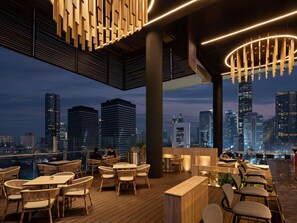 Un área de comedor al aire libre con mesas y sillas, una terraza de madera y vistas a un horizonte urbano.