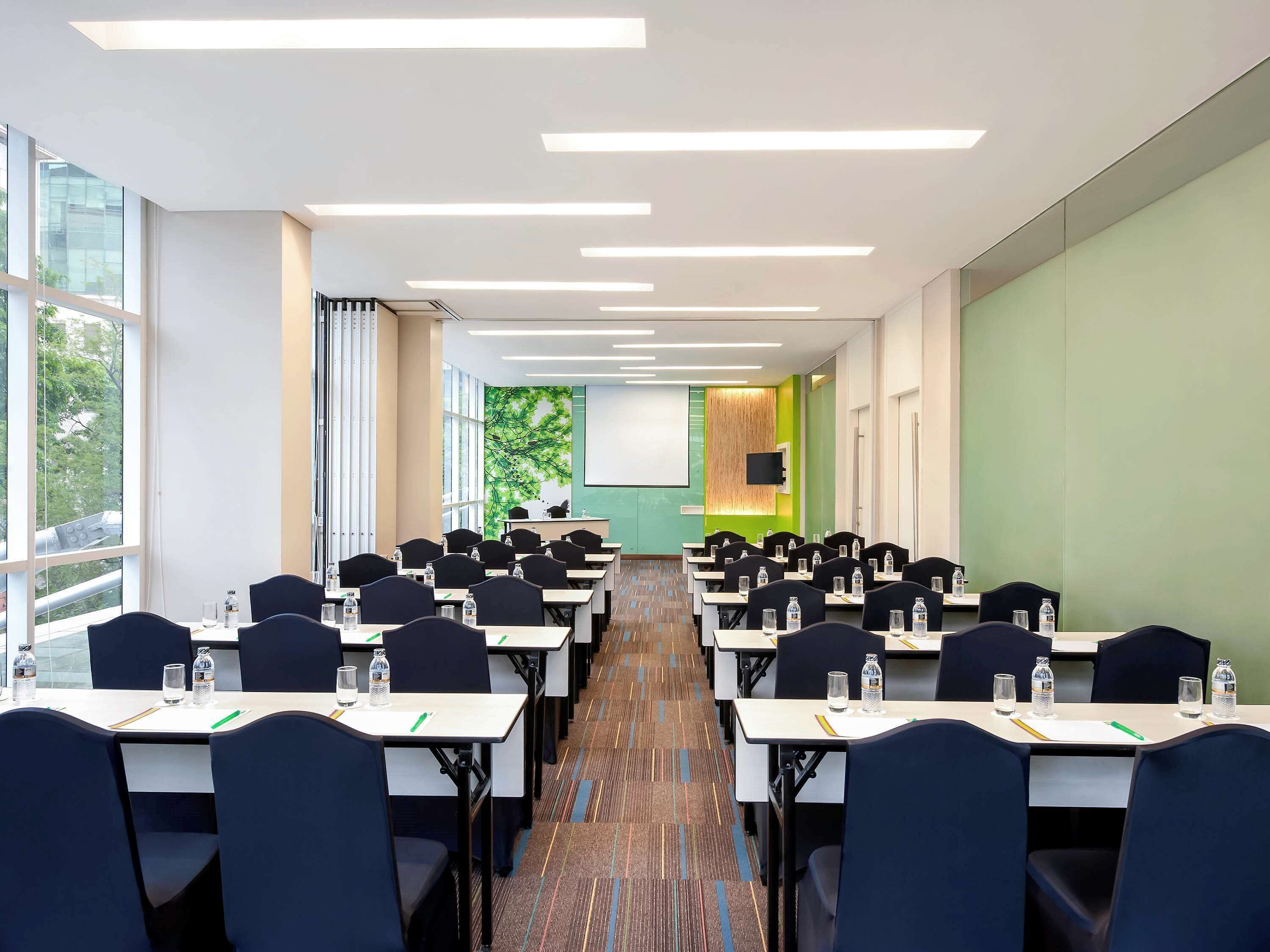 Una sala de conferències amb files de taules i cadires, una pantalla de projector i un gran finestral.
