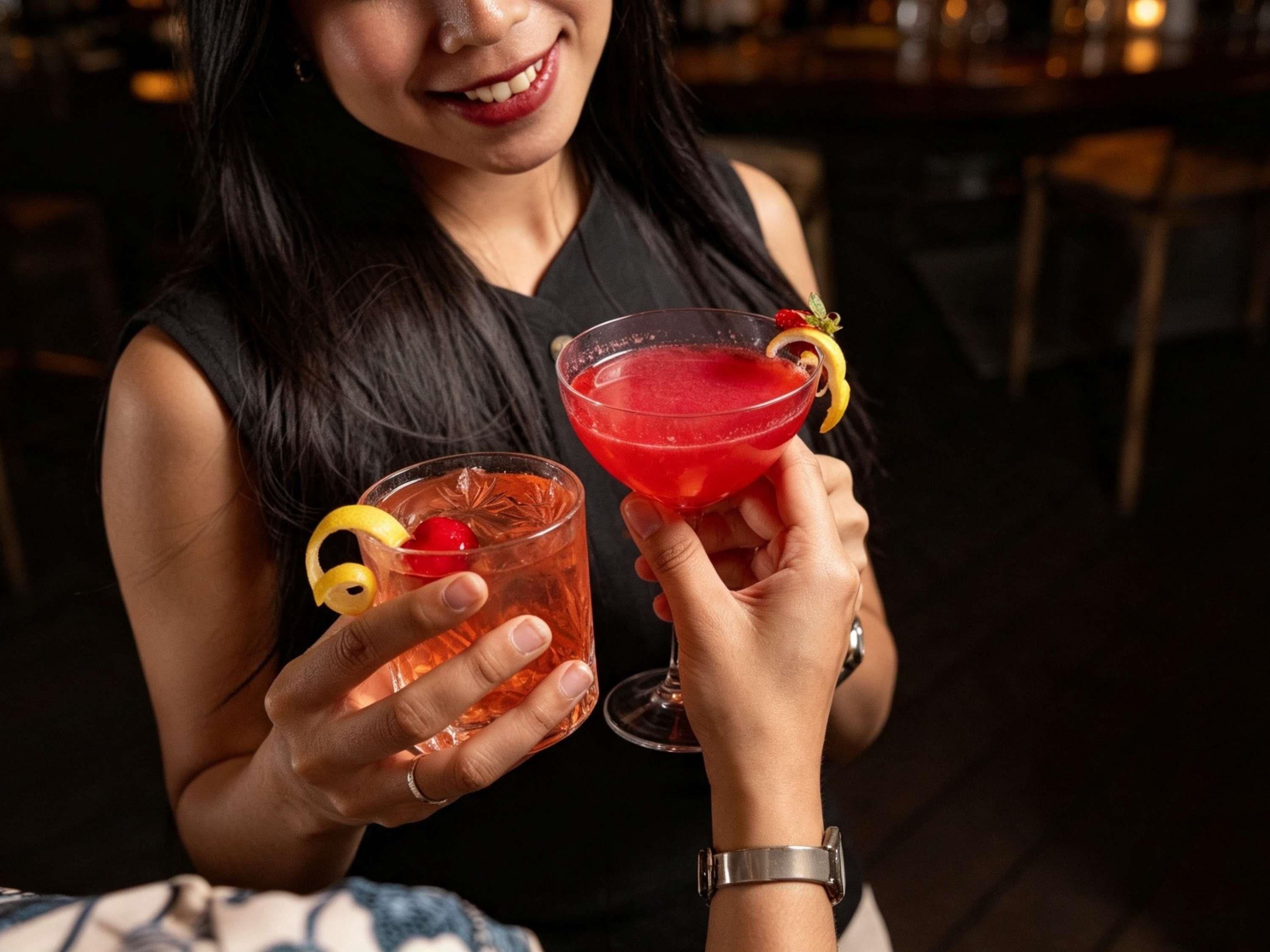 A person holding two glasses of a red beverage with garnishes, in a dimly lit indoor setting.