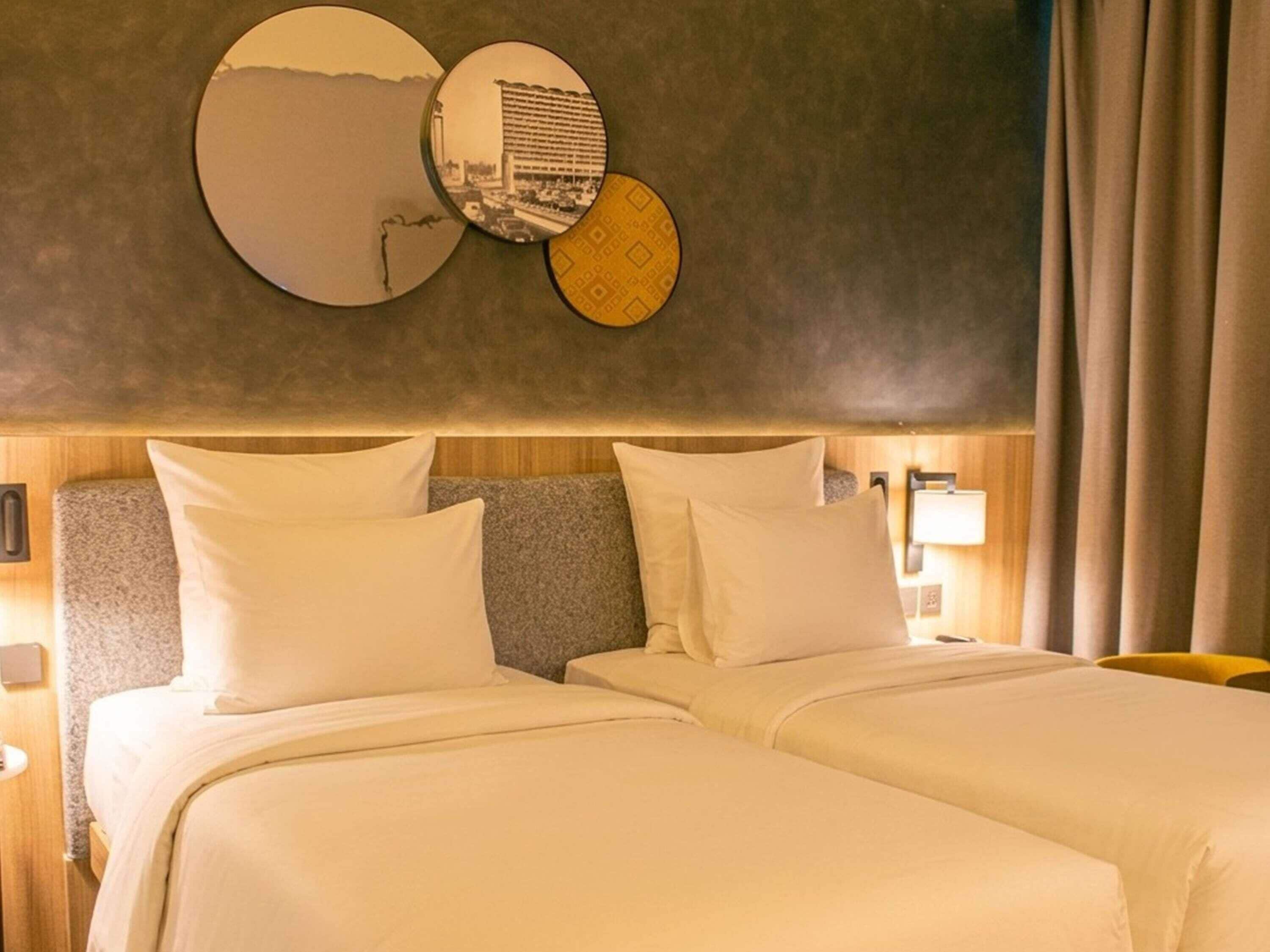 A hotel room with two beds, a large mirror, and a wall-mounted lamp.