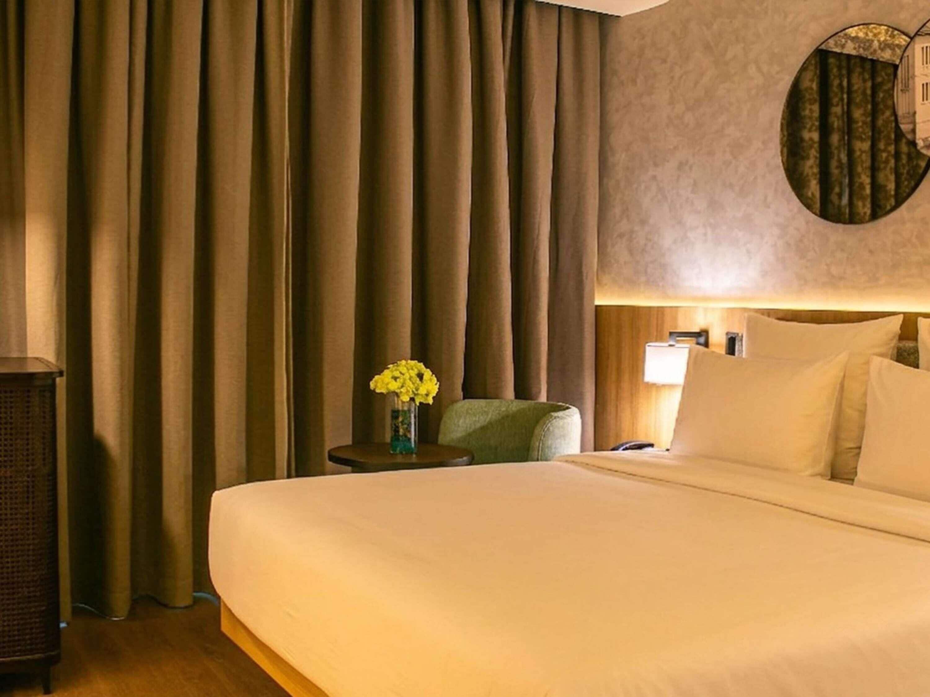 A hotel room with a large bed, a side table with a vase of flowers, a lamp, and a green chair.