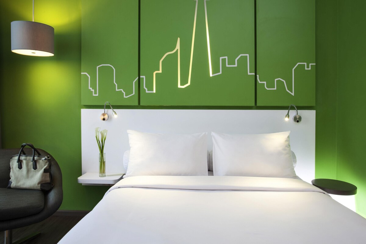A modern hotel room with a green accent wall, a city skyline light installation, a bed with white linens, a bedside table with a vase, and a chair with a handbag.