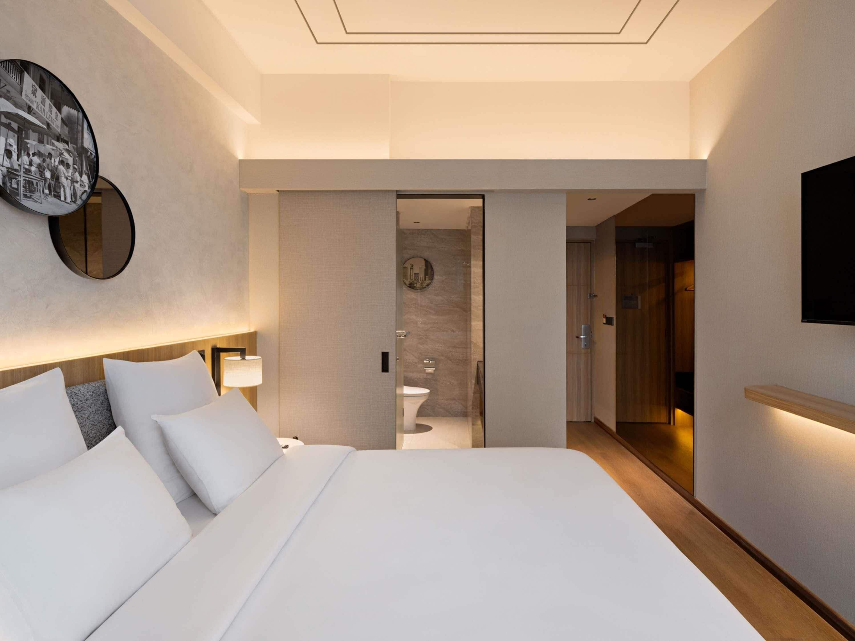 A modern hotel room with a large bed, a wall-mounted TV, and a bathroom visible through an open door.