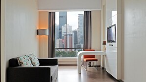 Une chambre d’hôtel moderne équipée d’un canapé, d’une table à manger avec une chaise, d’une télévision et d’une grande fenêtre donnant sur la ville.