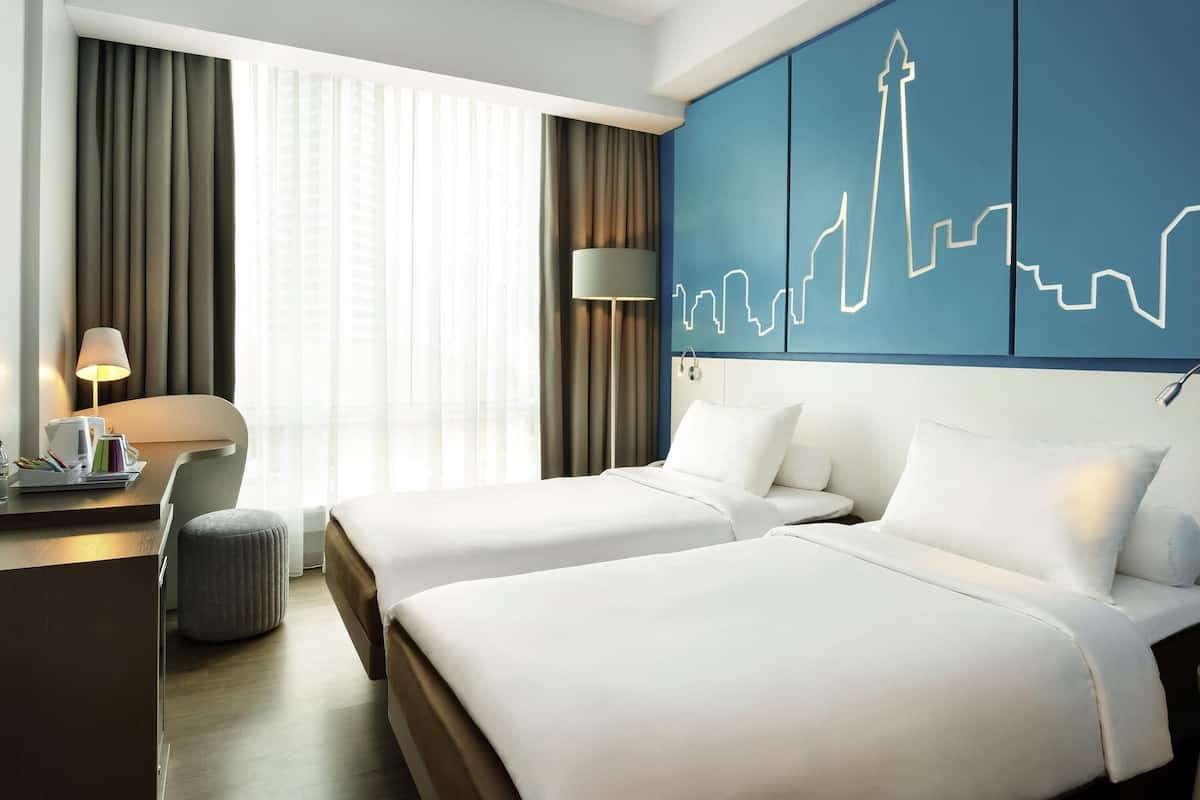 A hotel room with two beds, a desk with a lamp, a chair, and a large window with curtains.